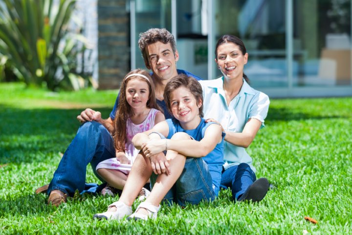 happy family sitting on grass in front of house, parents with two children smile