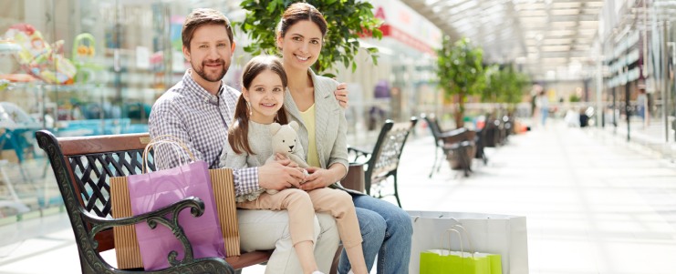 Adult parents with charming girl sitting on bench in big light shopping mall with colorful paper bags and smiling